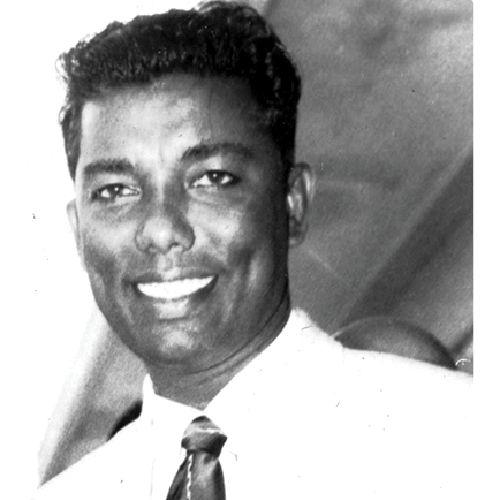 Black and white portrait of a man smiling, wearing a white shirt and patterned tie.