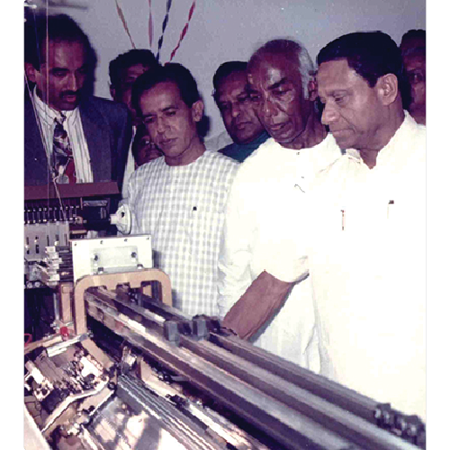 Group of men in formal and traditional attire observing a metallic mechanical device closely.