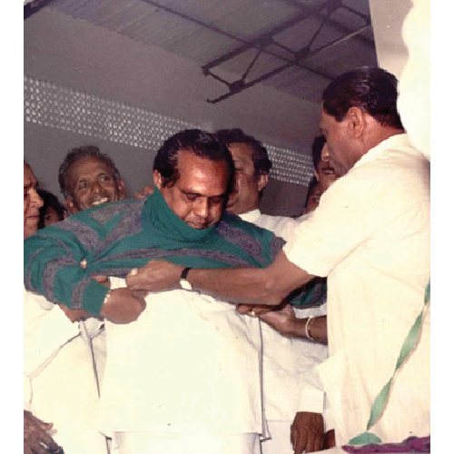 Group of men, with one helping another put on a white garment over his green and gray sweater indoors.