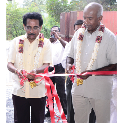 Two men wearing garlands cutting a red ribbon at an outdoor ceremony with two men in the background.