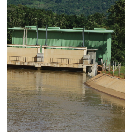 Green industrial building beside a water canal with surrounding greenery and hills in the background.