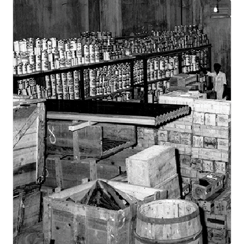 Black and white photo of a storage room filled with stacked cans on shelves, wooden crates, barrels, and a person standing near boxes in the back.