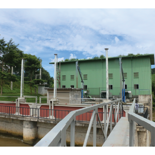 Small hydroelectric dam with green building and metal walkways surrounded by trees under a partly cloudy sky.