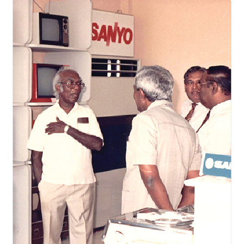 Four men in formal and casual attire engaged in conversation inside a showroom with Sanyo brand electronics displayed.