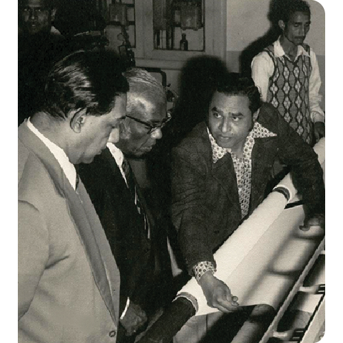 Three men closely examining a large roll of fabric on a machine, with a fourth man standing in the background.