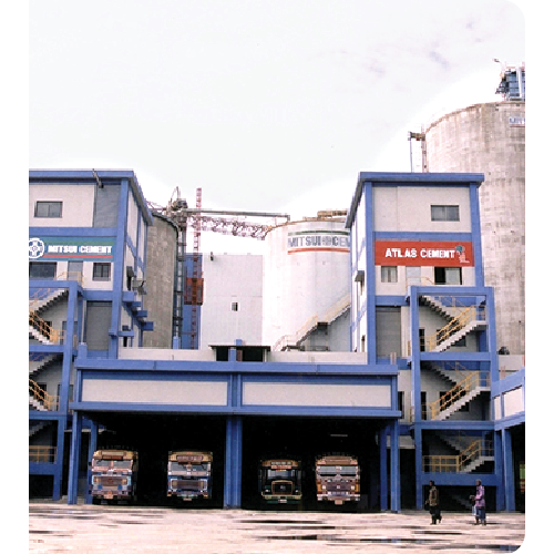 Industrial cement plant with blue and gray buildings labeled Mitsui Cement and Atlas Cement, with trucks parked underneath and two people walking nearby.