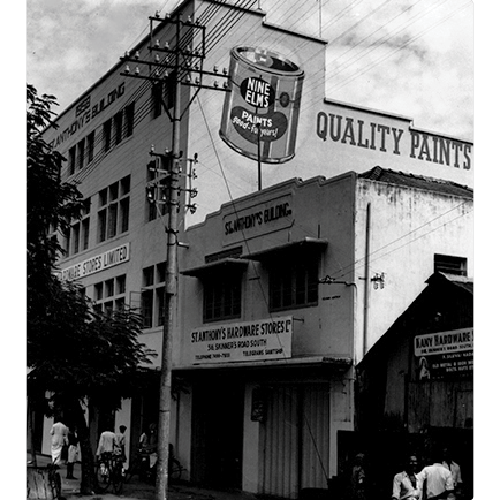 Black and white photo of a street corner with buildings advertising hardware and paint stores, featuring a large floating paint can sign that reads 'NINE ELMS PAINTS'.