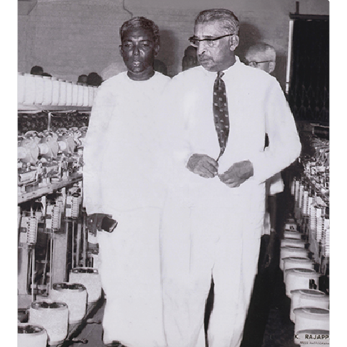 Two men walking between rows of yarn spools and textile machinery in a factory setting.
