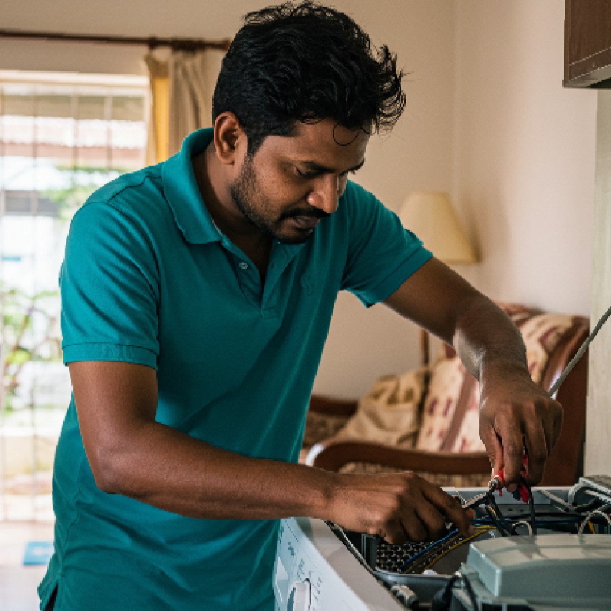 Man in teal shirt repairing electrical appliance with screwdriver inside a home.